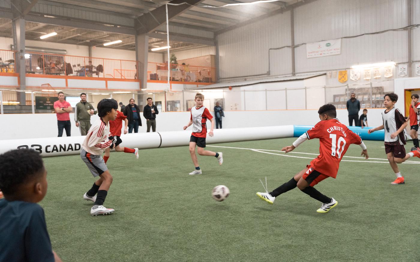 Young players in a fast 3v3 match inside an AirPitch, the inflatable boundary keeping the ball in play as a player strikes toward goal.