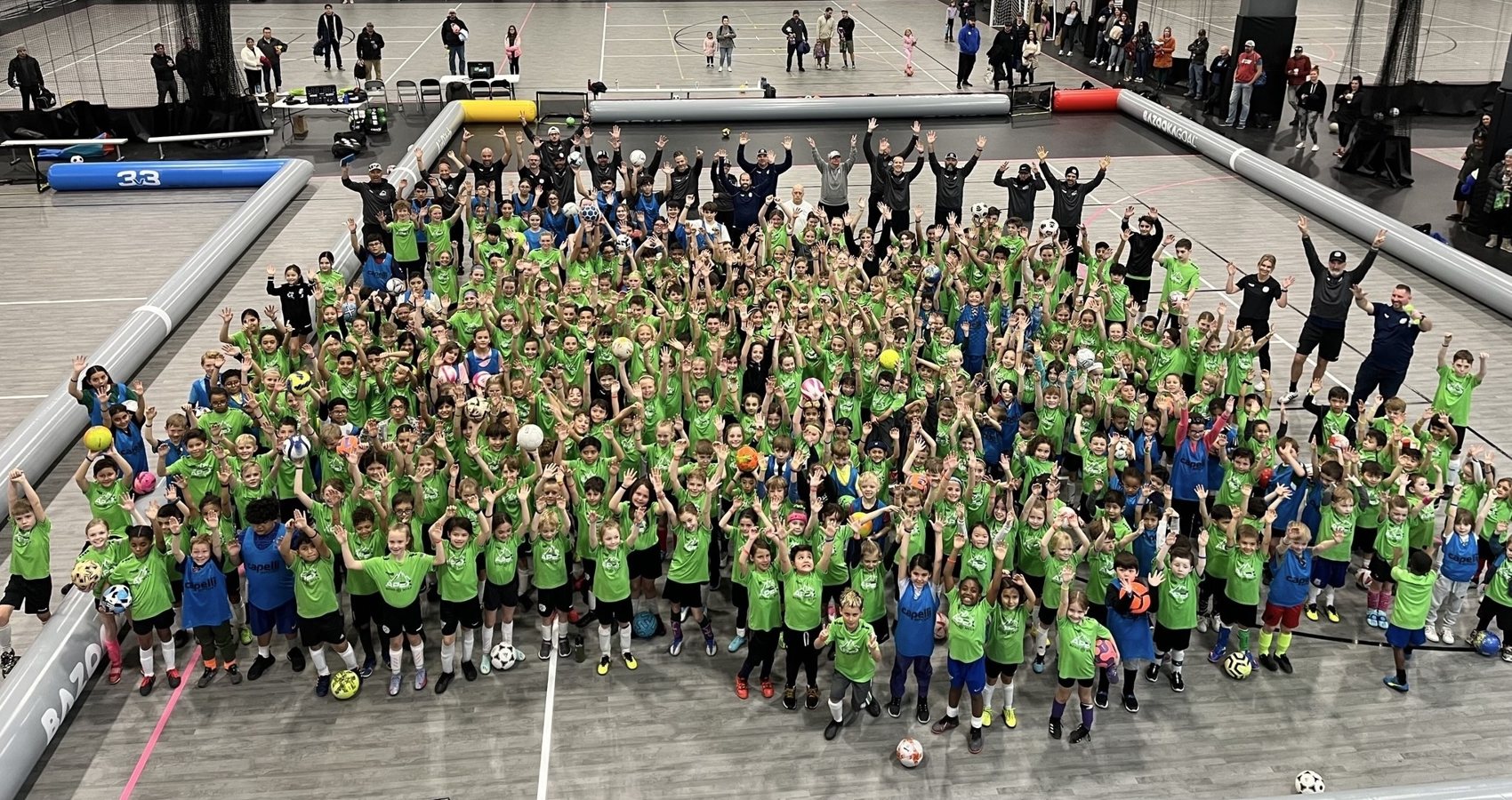 Hundreds of young players in 3v3 jerseys with arms raised around the AirPitch at an indoor event in Winnipeg.