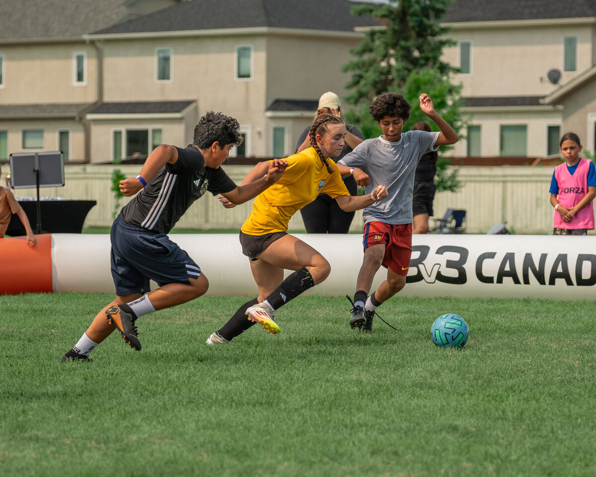 Three players chasing the ball during a match, with the 3v3 Canada AirPitch boundary behind them.
